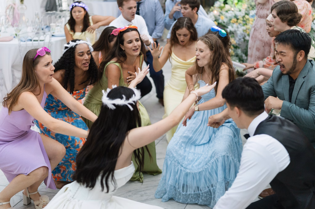 Robbie & Hannah's Wedding @ Edinburgh West, Taylors SC: Wedding guests in colorful outfits crouch and dance together indoors, wearing playful light-up sunglasses and feather accessories.
