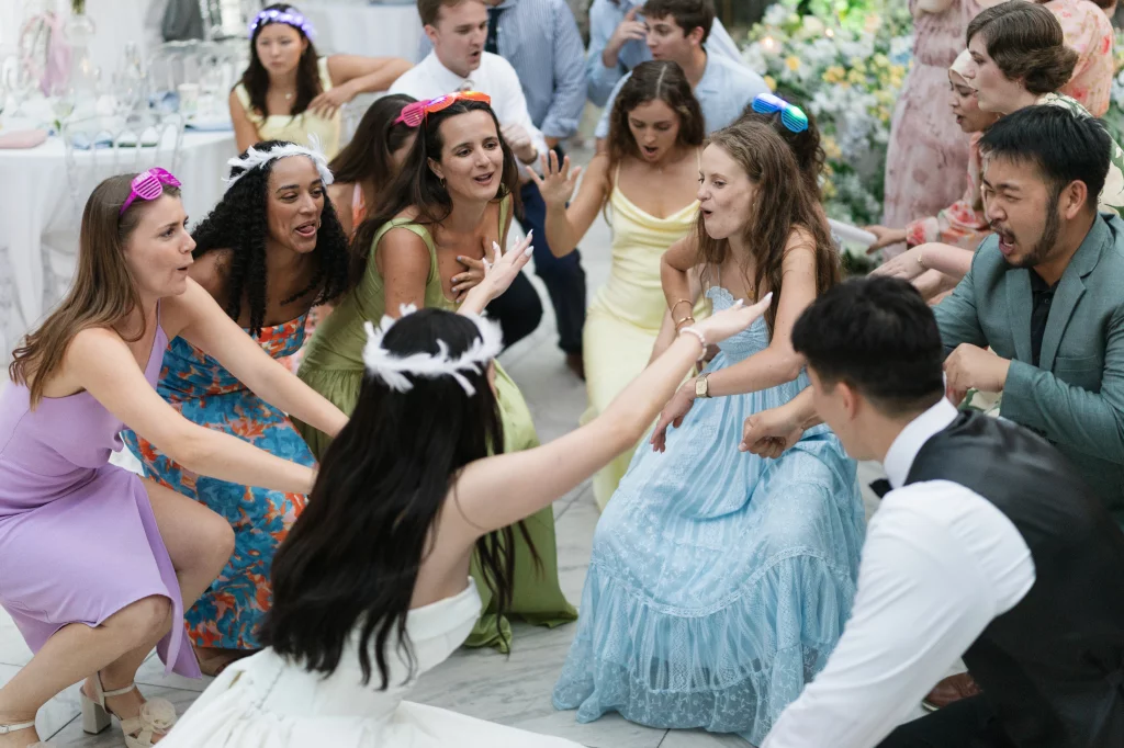 Robbie & Hannah's Wedding @ Edinburgh West, Taylors SC: Wedding guests in colorful outfits crouch and dance together indoors, wearing playful light-up sunglasses and feather accessories.