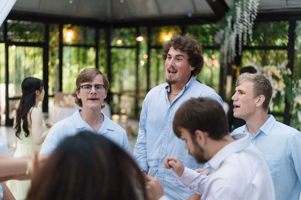 Robbie & Hannah's Wedding @ Edinburgh West, Taylors SC: Wedding guests in light shirts chat inside a glass-walled venue decorated with greenery and flowers.