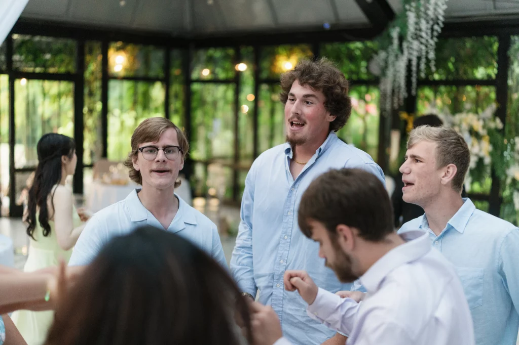 Robbie & Hannah's Wedding @ Edinburgh West, Taylors SC: Wedding guests in light shirts chat inside a glass-walled venue decorated with greenery and flowers.