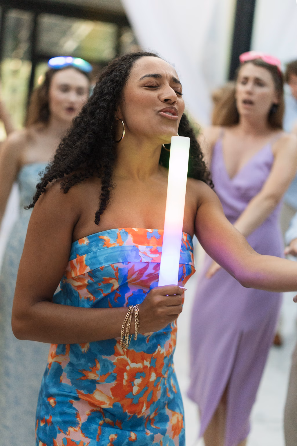 Robbie & Hannah's Wedding @ Edinburgh West, Taylors SC: Woman in a colorful floral dress holds a glowing foam stick while dancing, with wedding guests behind her.