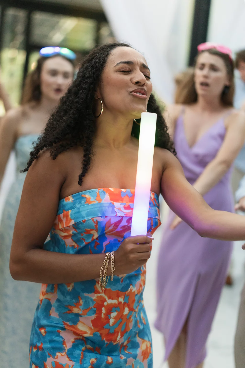 Robbie & Hannah's Wedding @ Edinburgh West, Taylors SC: Woman in a colorful floral dress holds a glowing foam stick while dancing, with wedding guests behind her.