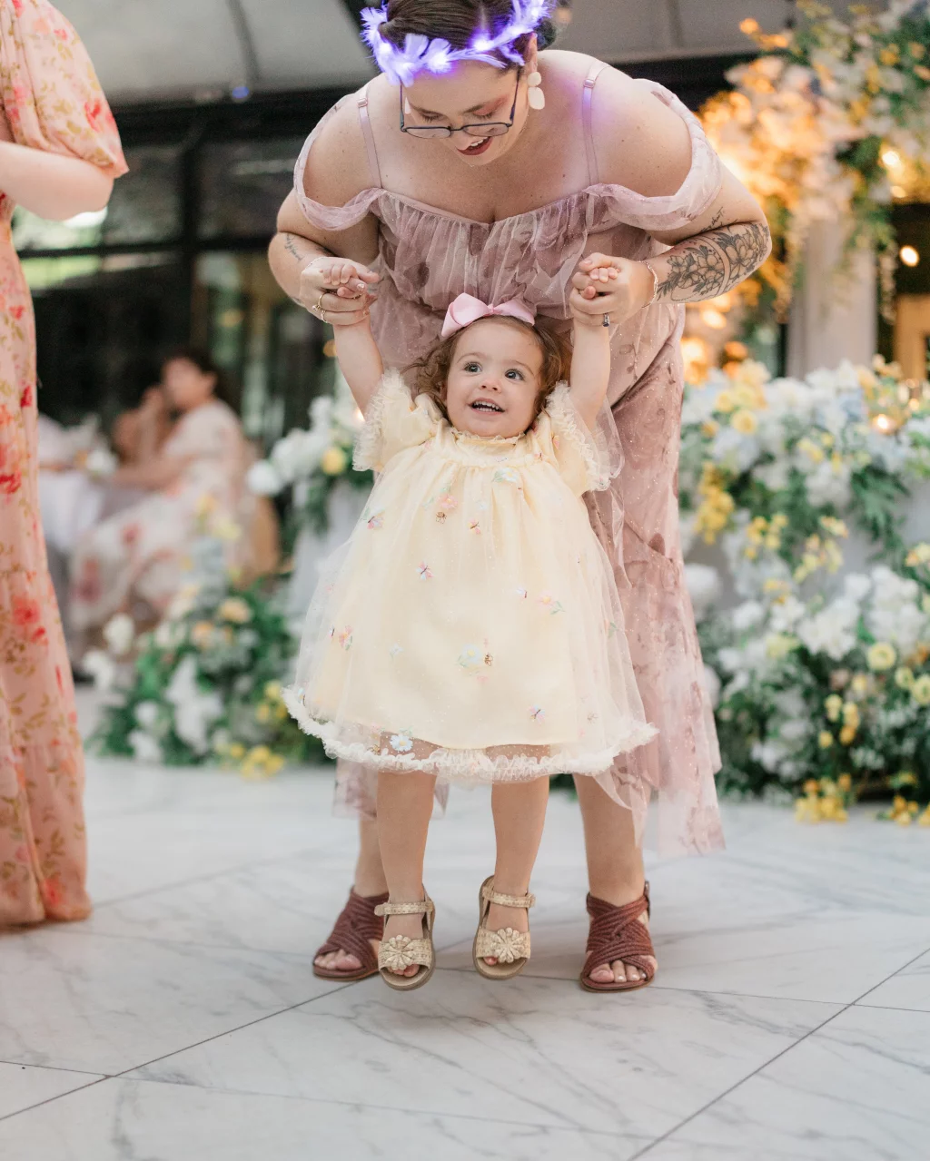 Robbie & Hannah's Wedding @ Edinburgh West, Taylors SC: Woman in a pink dress holds a smiling toddler in a floral dress during a flower-filled wedding celebration.