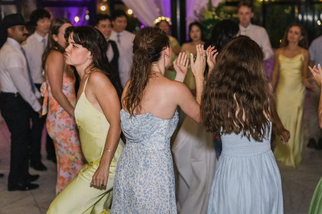 Robbie & Hannah's Wedding @ Edinburgh West, Taylors SC: Wedding guests dance together indoors under purple lighting, with three women in pastel dresses in the foreground.