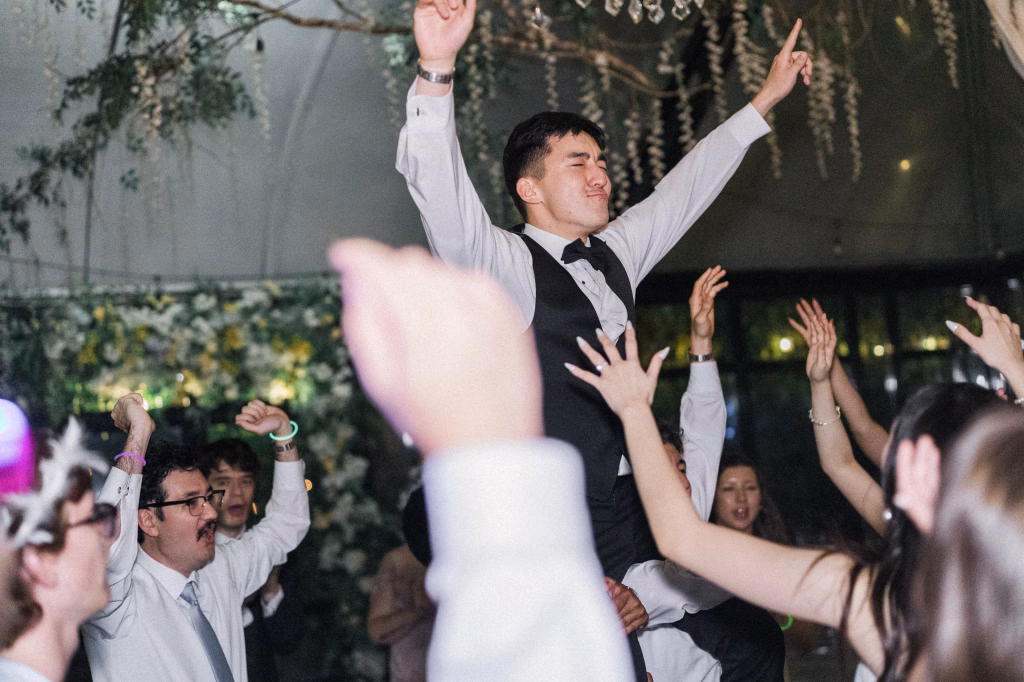 Robbie & Hannah's Wedding @ Edinburgh West, Taylors SC: Wedding guests cheer and lift a smiling man in a vest and bow tie above the crowd.