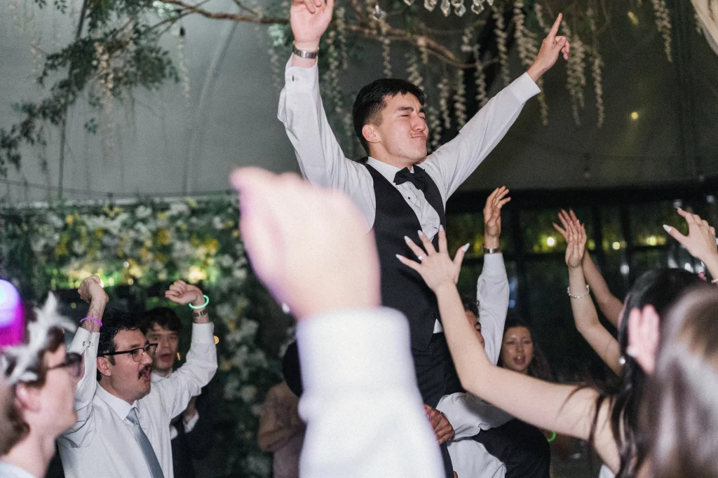 Robbie & Hannah's Wedding @ Edinburgh West, Taylors SC: Wedding guests cheer and lift a smiling man in a vest and bow tie above the crowd.