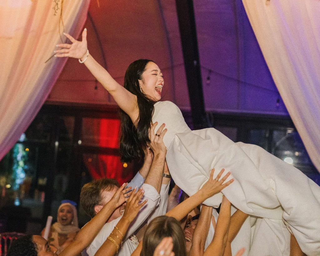 Robbie & Hannah's Wedding @ Edinburgh West, Taylors SC: Bride in a white dress is lifted joyfully by wedding guests beneath colorful draped lights.