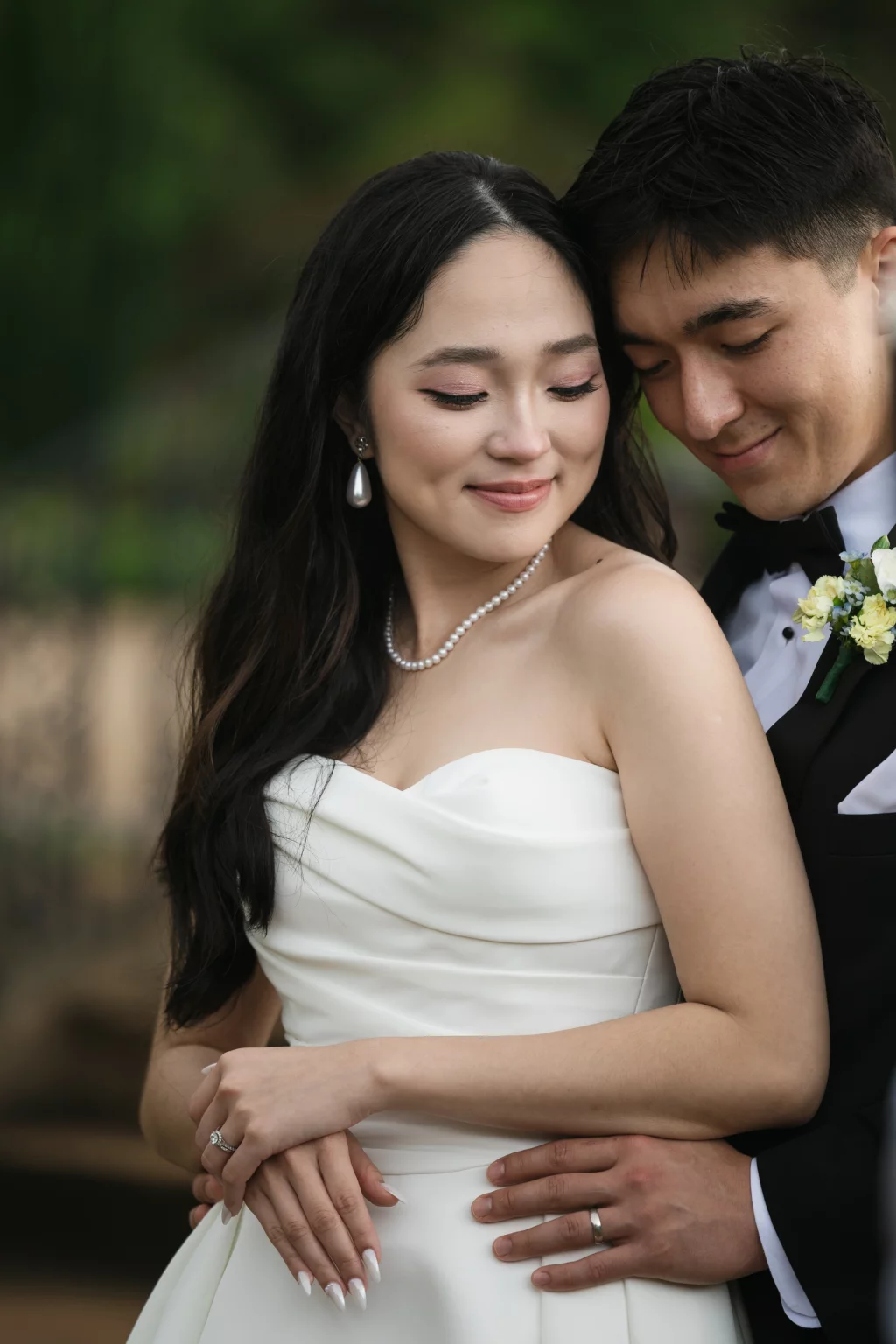 Robbie & Hannah's Wedding @ Edinburgh West, Taylors SC: Bride in a strapless white gown and pearl necklace embraces her groom in a black tuxedo, both smiling at their wedding.
