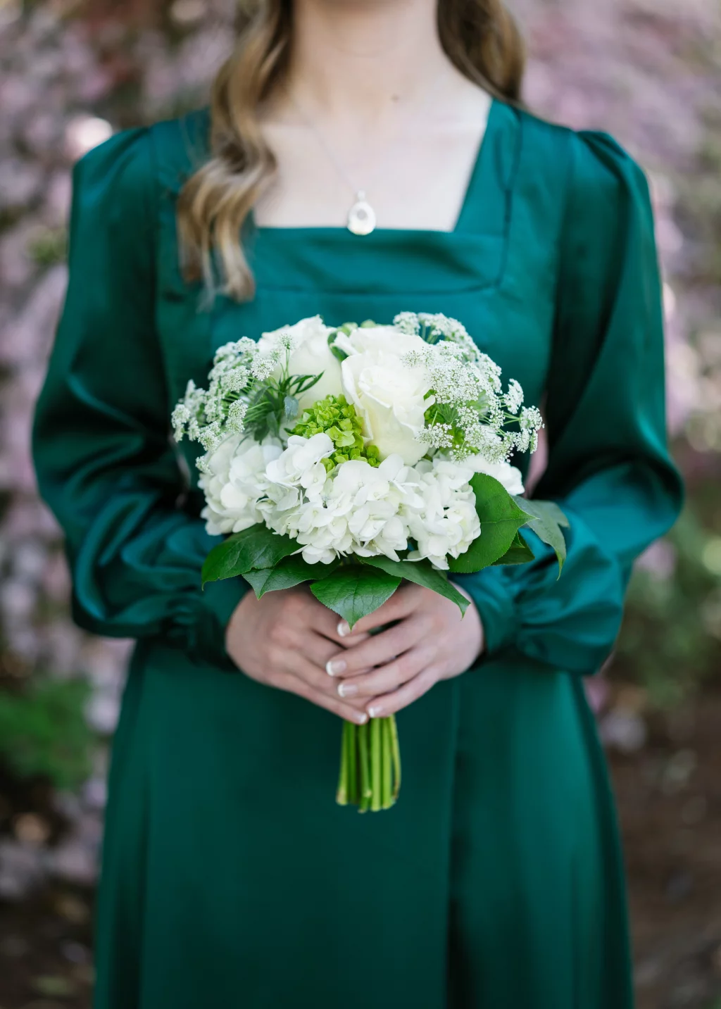 BJA Jr/Sr Banquet Prom at Sue Simpson Park: A woman in a dark green dress holds a bouquet of white flowers and greenery, standing outdoors with a blurred floral background.