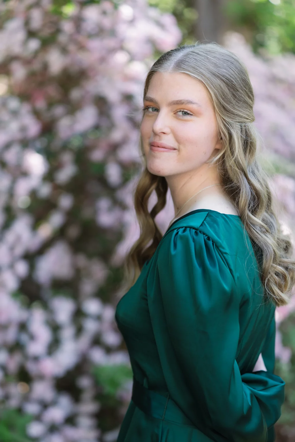 BJA Jr/Sr Banquet Prom at Sue Simpson Park: A young woman with long, wavy blonde hair wearing a green dress stands outdoors, smiling softly with pink flowers blurred in the background.