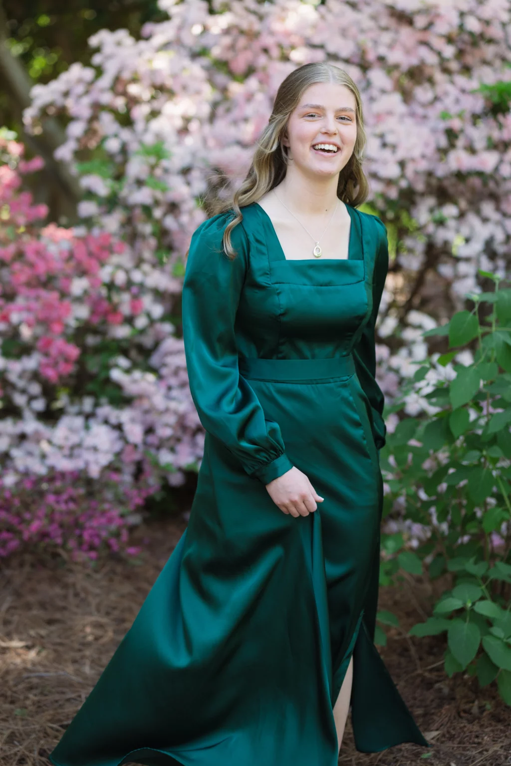 BJA Jr/Sr Banquet Prom at Sue Simpson Park: Young woman in a long, emerald green dress standing outdoors, smiling in front of pink and white flowering bushes.