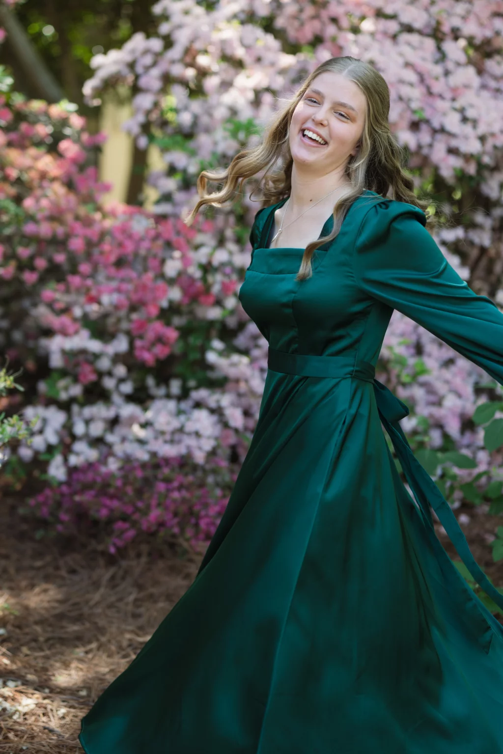 BJA Jr/Sr Banquet Prom at Sue Simpson Park: Smiling young woman in a long green dress twirls outdoors in front of blooming pink and white flowers.