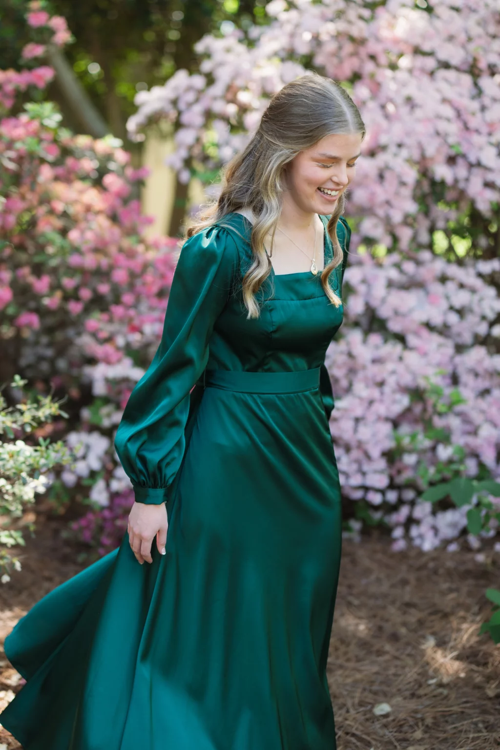 BJA Jr/Sr Banquet Prom at Sue Simpson Park: Young woman in a long, emerald green dress walking outdoors, smiling, with blooming pink flowers in the background.