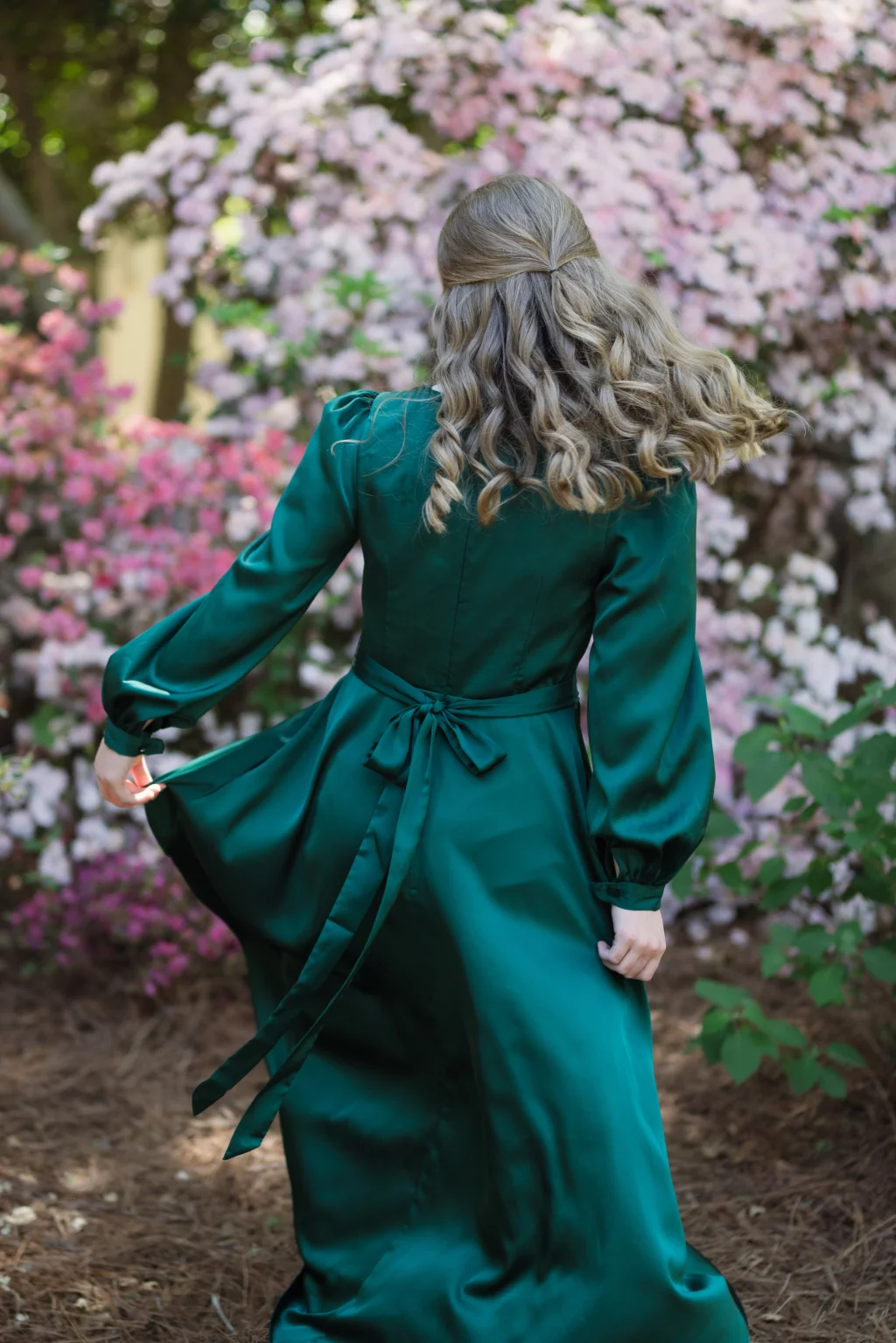 BJA Jr/Sr Banquet Prom at Sue Simpson Park: A person with wavy blonde hair in a green satin dress stands outdoors, facing away, surrounded by blooming pink and white flowers.