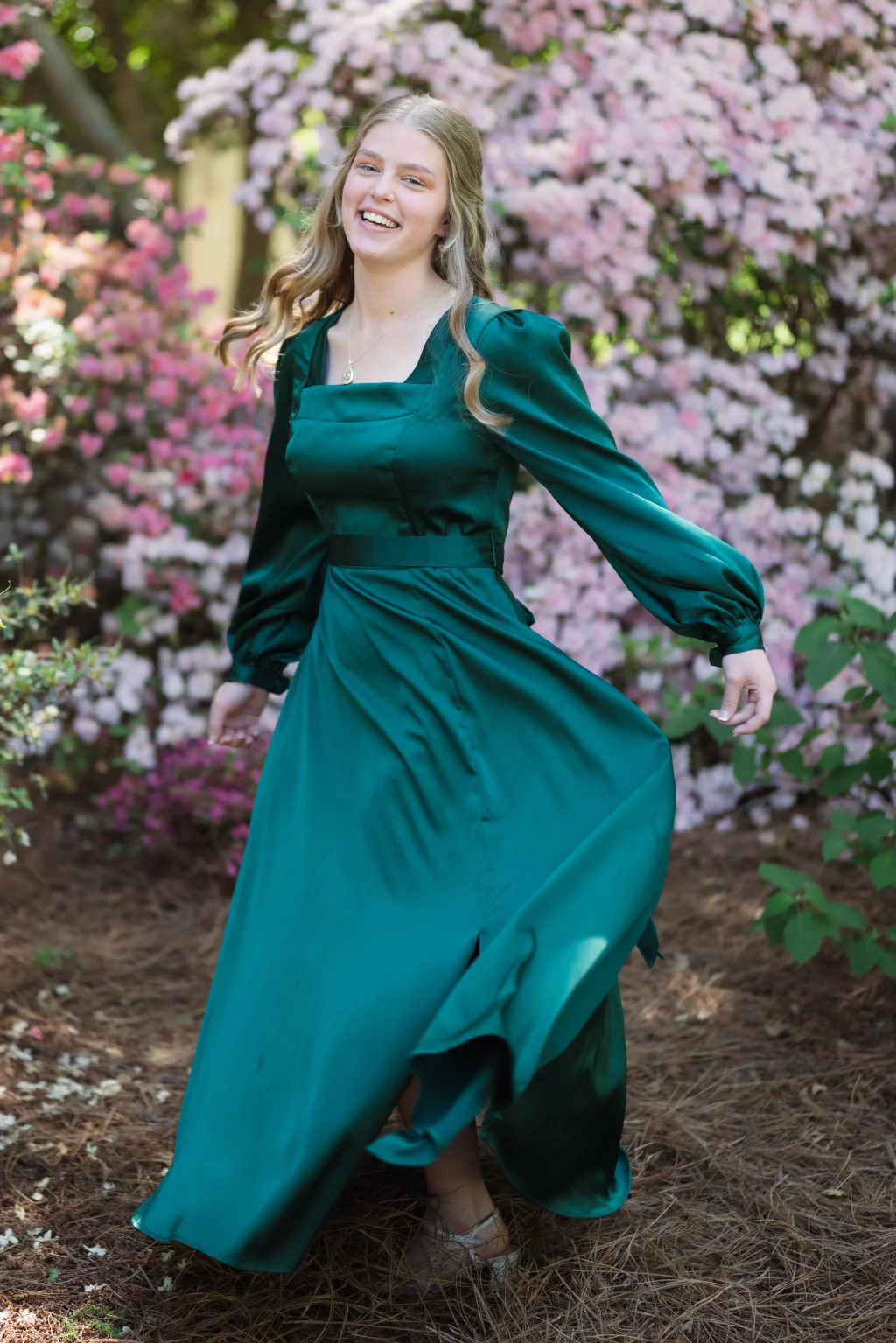 BJA Jr/Sr Banquet Prom at Sue Simpson Park: Young woman in a long, flowing emerald green dress smiling and twirling outdoors in front of blooming pink flowers.