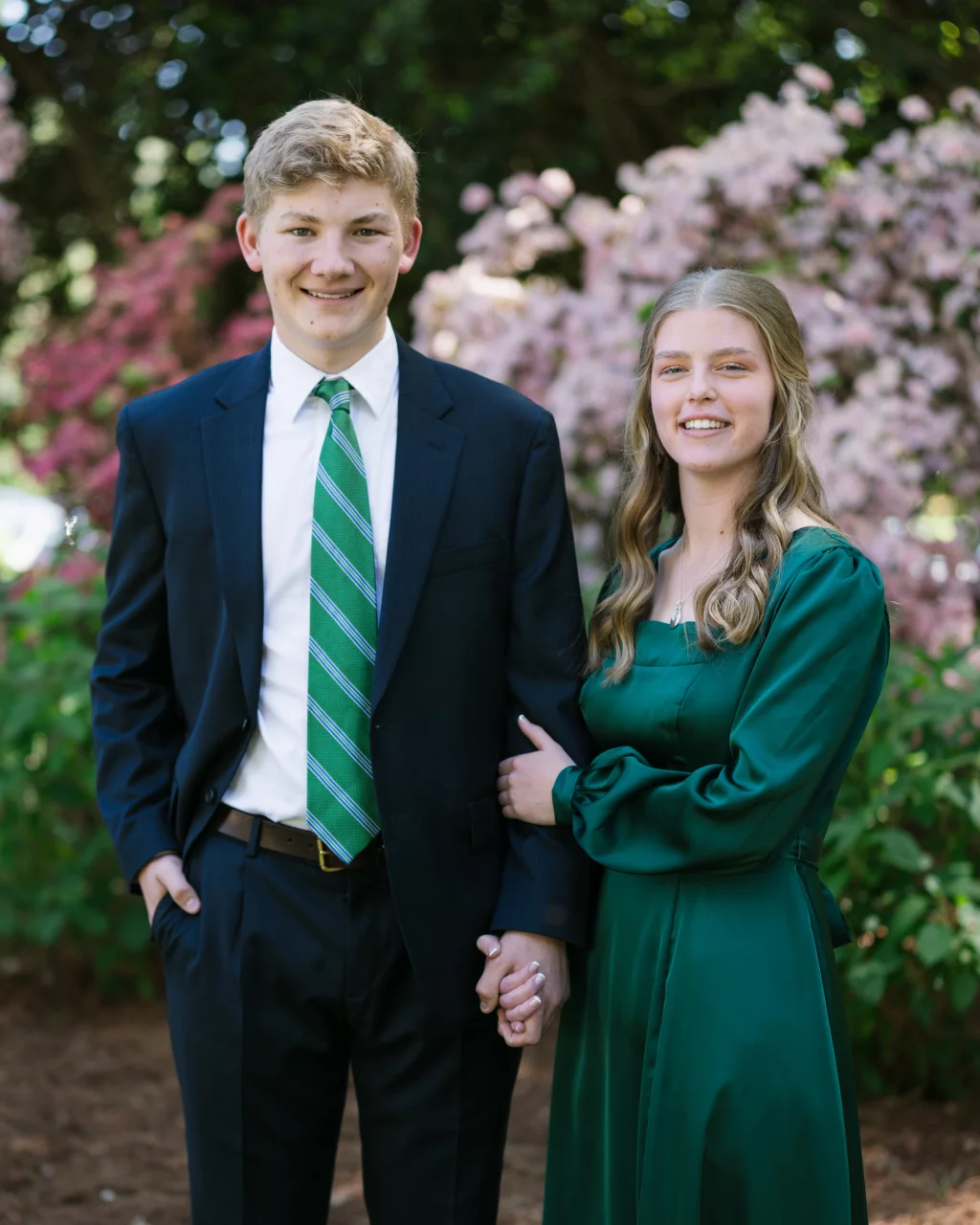 BJA Jr/Sr Banquet Prom at Sue Simpson Park: A young man in a dark suit with a green striped tie stands holding hands with a young woman in a long green dress. They are smiling and posing outdoors in front of blooming pink flowers and greenery.