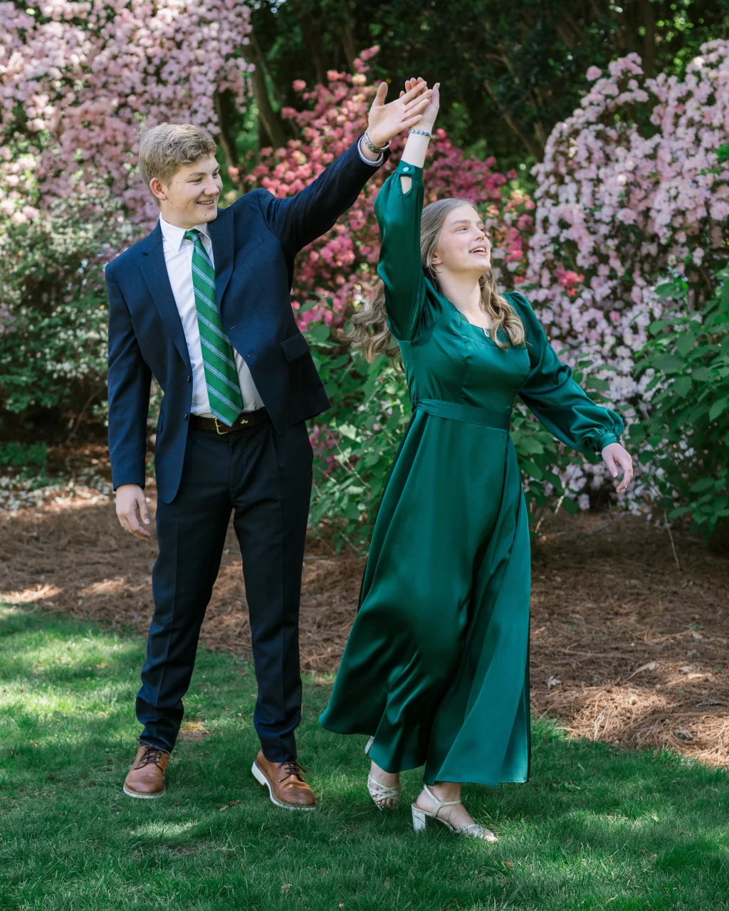 BJA Jr/Sr Banquet Prom at Sue Simpson Park: A young man in a suit and green striped tie twirls a young woman in a long green dress on a grassy lawn, with blooming pink bushes in the background.