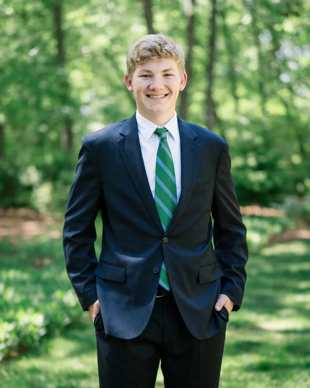 BJA Jr/Sr Banquet Prom at Sue Simpson Park: Young man in a dark suit with a green striped tie, standing outdoors in a sunlit, wooded area, smiling with hands in pockets.