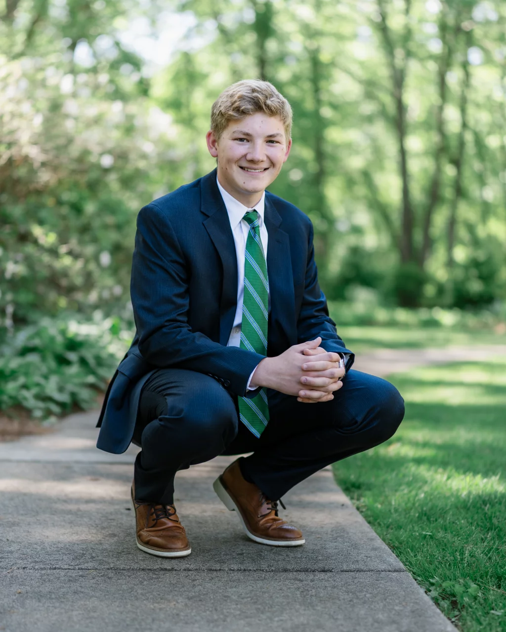 BJA Jr/Sr Banquet Prom at Sue Simpson Park: Young man with blond hair wearing a navy suit, white shirt, and green striped tie, squatting and smiling on a sidewalk in a green, sunlit park.