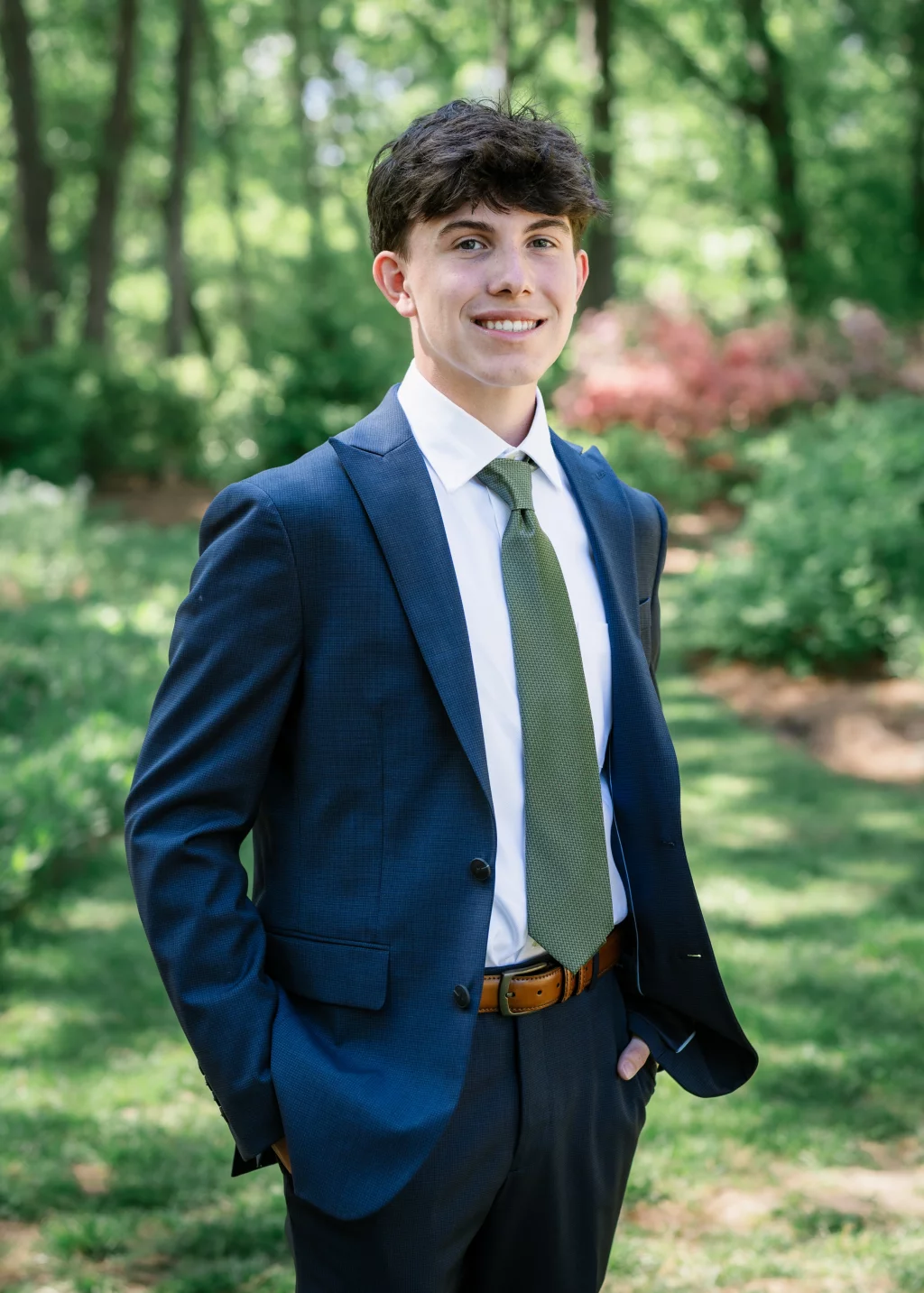 BJA Jr/Sr Banquet Prom at Sue Simpson Park: Young man in a blue suit, white shirt, and green tie standing outdoors in a green, sunlit garden with trees and bushes in the background.