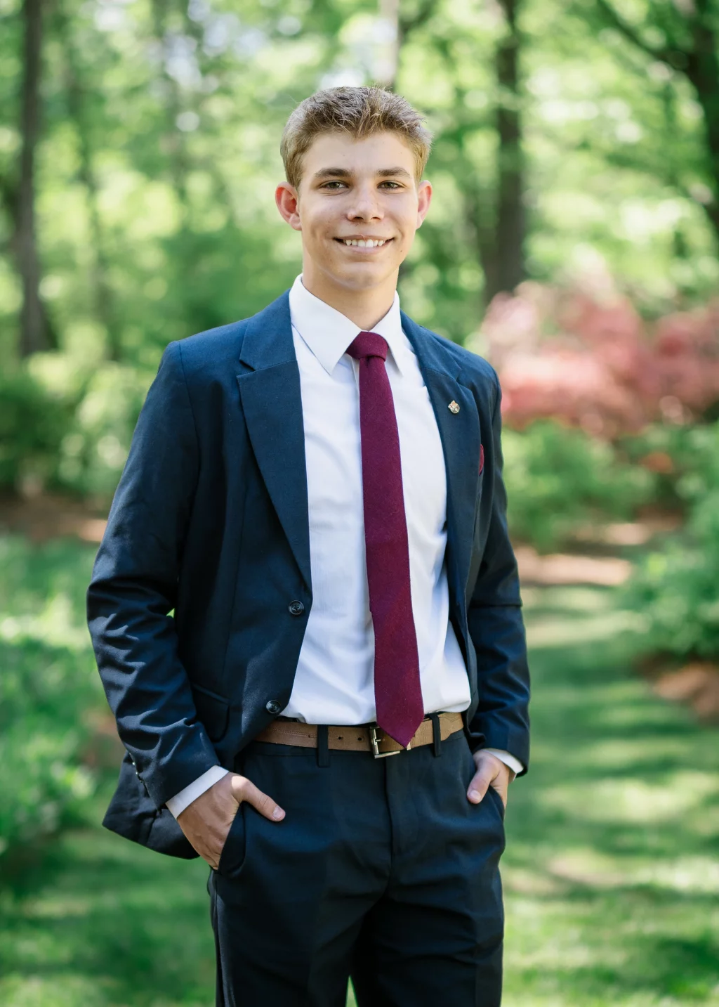 BJA Jr/Sr Banquet Prom at Sue Simpson Park: Young man in a navy suit, white shirt, and maroon tie standing outdoors in a sunlit, green garden, smiling with hands in his pockets.