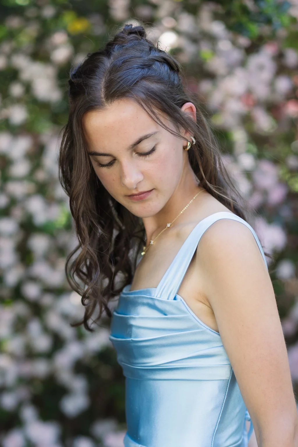 BJA Jr/Sr Banquet Prom at Sue Simpson Park: Young woman with long brown hair in a light blue satin dress, looking down with eyes closed, standing outdoors in front of a blurred background of pink flowers and greenery.
