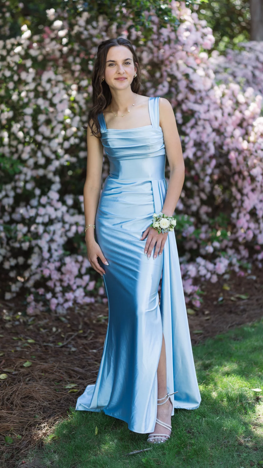 BJA Jr/Sr Banquet Prom at Sue Simpson Park: Young woman in a shiny light blue formal gown with a white corsage on her wrist, standing outdoors in front of blooming pink flowers.