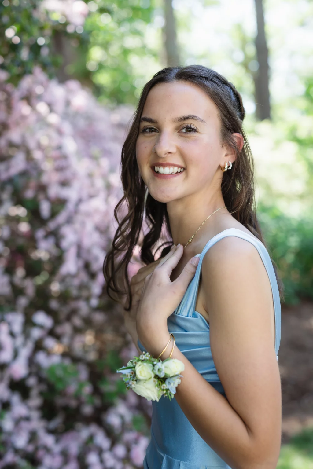 BJA Jr/Sr Banquet Prom at Sue Simpson Park: Young woman in a light blue dress smiling outdoors, wearing a white floral corsage on her wrist, with blooming pink flowers and greenery in the background.