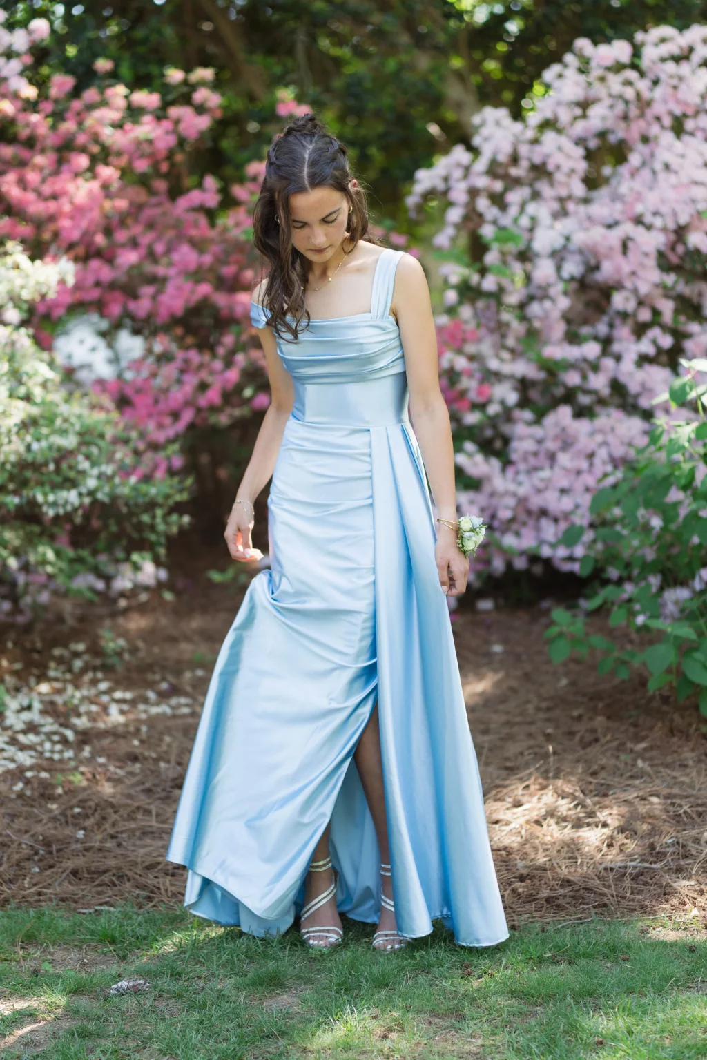 BJA Jr/Sr Banquet Prom at Sue Simpson Park: Young woman in a light blue satin gown standing outdoors, surrounded by blooming pink and white flowers, looking down and holding her dress.