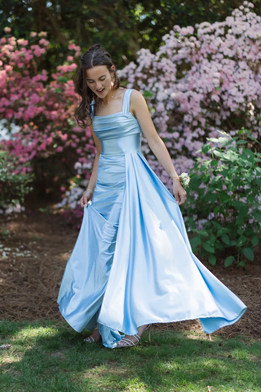 BJA Jr/Sr Banquet Prom at Sue Simpson Park: A young woman in a long, flowing light blue gown stands outdoors, holding the skirt of her dress. She has long brown hair and is wearing a white corsage on her wrist. Pink and white flowering bushes are in the background.