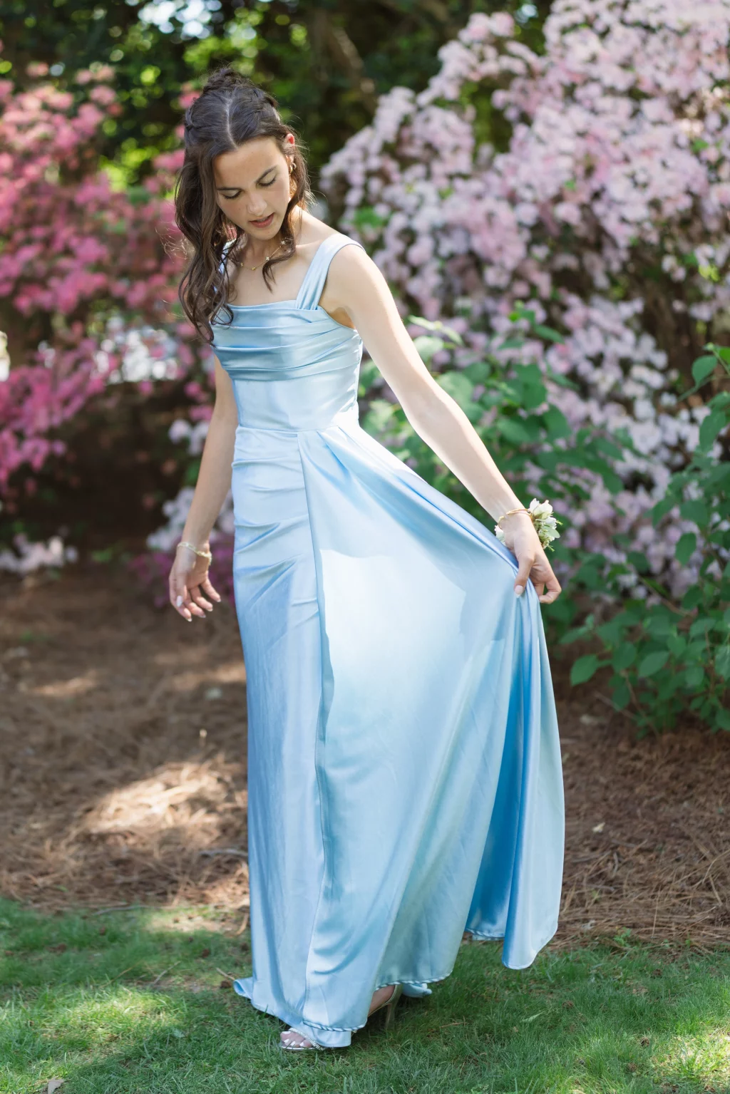 BJA Jr/Sr Banquet Prom at Sue Simpson Park: Young woman in a light blue satin gown standing outdoors, holding one side of her dress, with blooming pink and white flowers in the background.