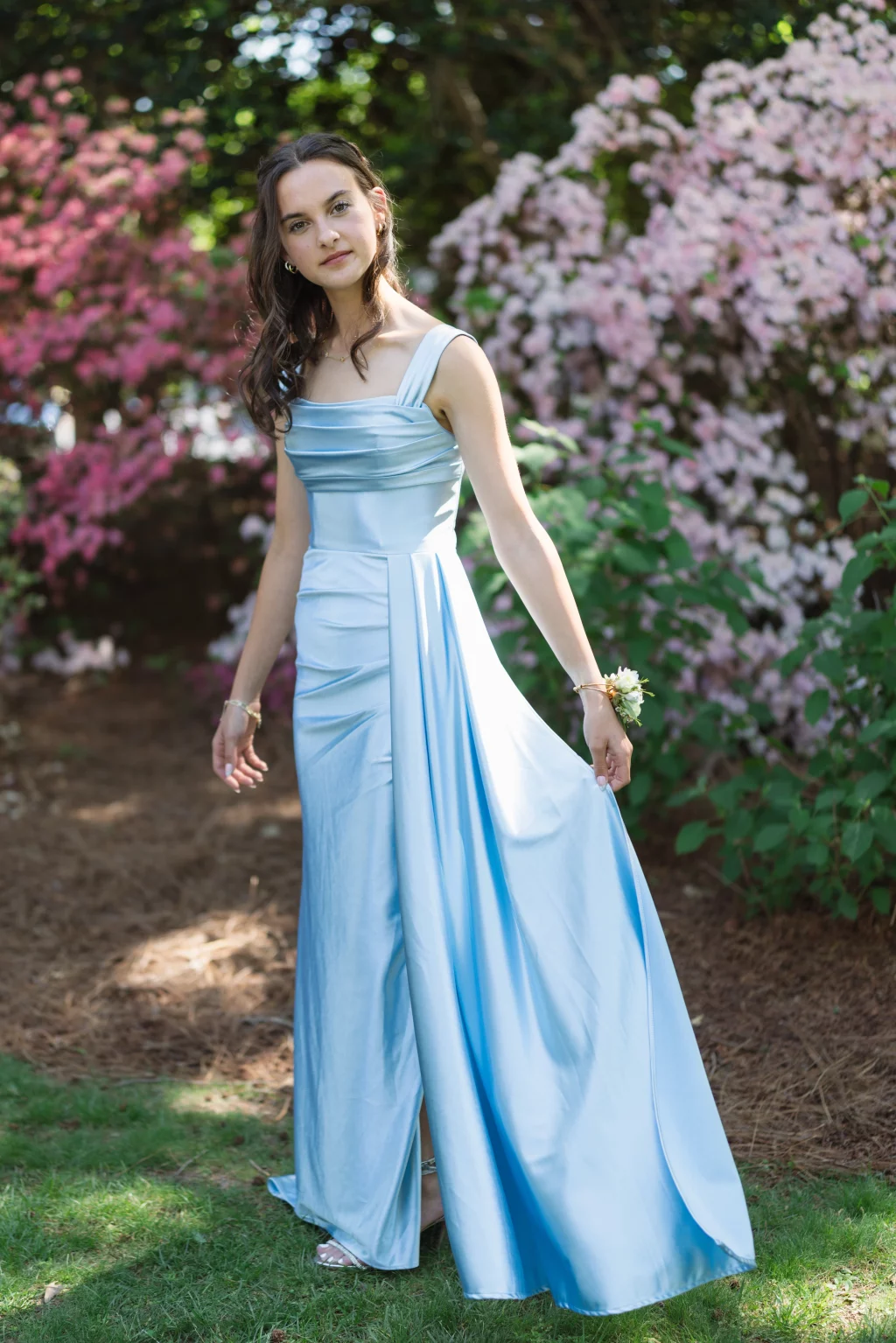 BJA Jr/Sr Banquet Prom at Sue Simpson Park: Young woman in a light blue formal gown standing on grass, holding the skirt, with pink and white flowering bushes in the background.
