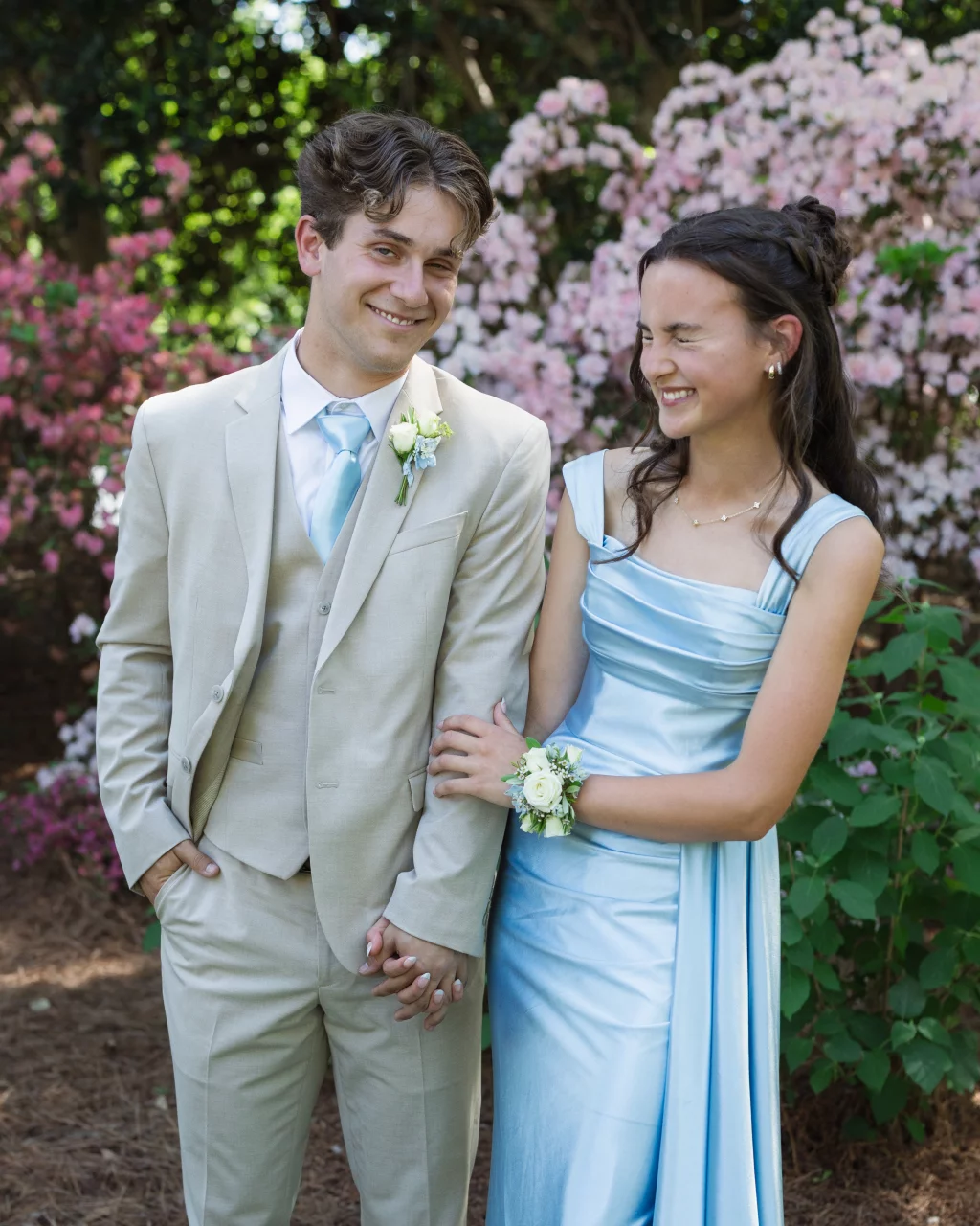 BJA Jr/Sr Banquet Prom at Sue Simpson Park: A young couple dressed for a formal event stands smiling in front of pink flowering bushes; the young man wears a beige suit with a light blue tie and boutonniere, while the young woman wears a shiny light blue gown and corsage, linking arms and holding hands.