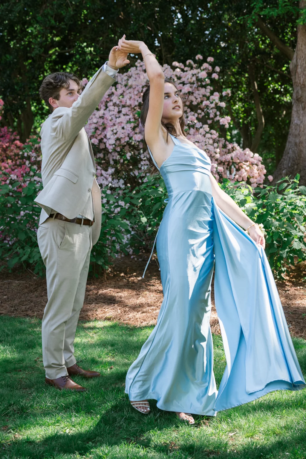 BJA Jr/Sr Banquet Prom at Sue Simpson Park: A young man in a light suit twirls a young woman in a long, light blue satin dress in a garden with pink flowering bushes.