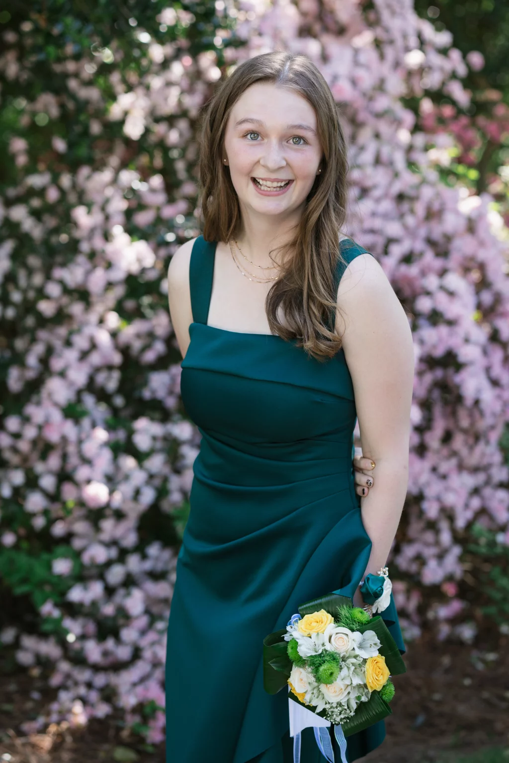 BJA Jr/Sr Banquet Prom at Sue Simpson Park: Young woman in a dark green dress holding a small bouquet of yellow, white, and green flowers, standing in front of pink flowering bushes and smiling at the camera.