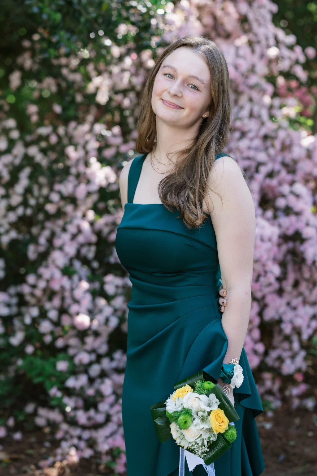 BJA Jr/Sr Banquet Prom at Sue Simpson Park: Young woman in a dark teal dress holding a bouquet of yellow, white, and green flowers, standing in front of blooming pink bushes.