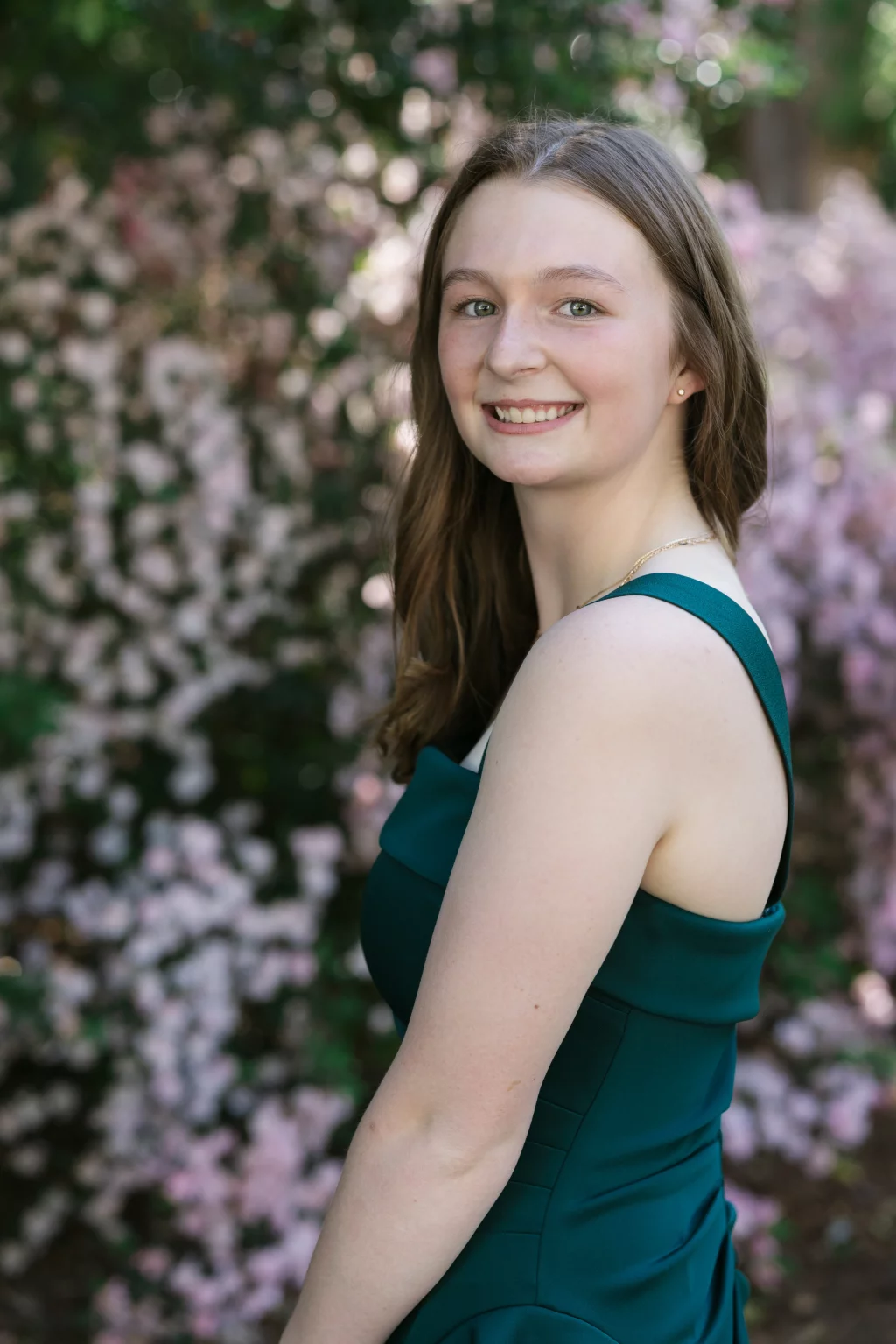 BJA Jr/Sr Banquet Prom at Sue Simpson Park: A young woman with long brown hair wearing a sleeveless teal dress smiles while standing outdoors in front of blooming pink flowers.