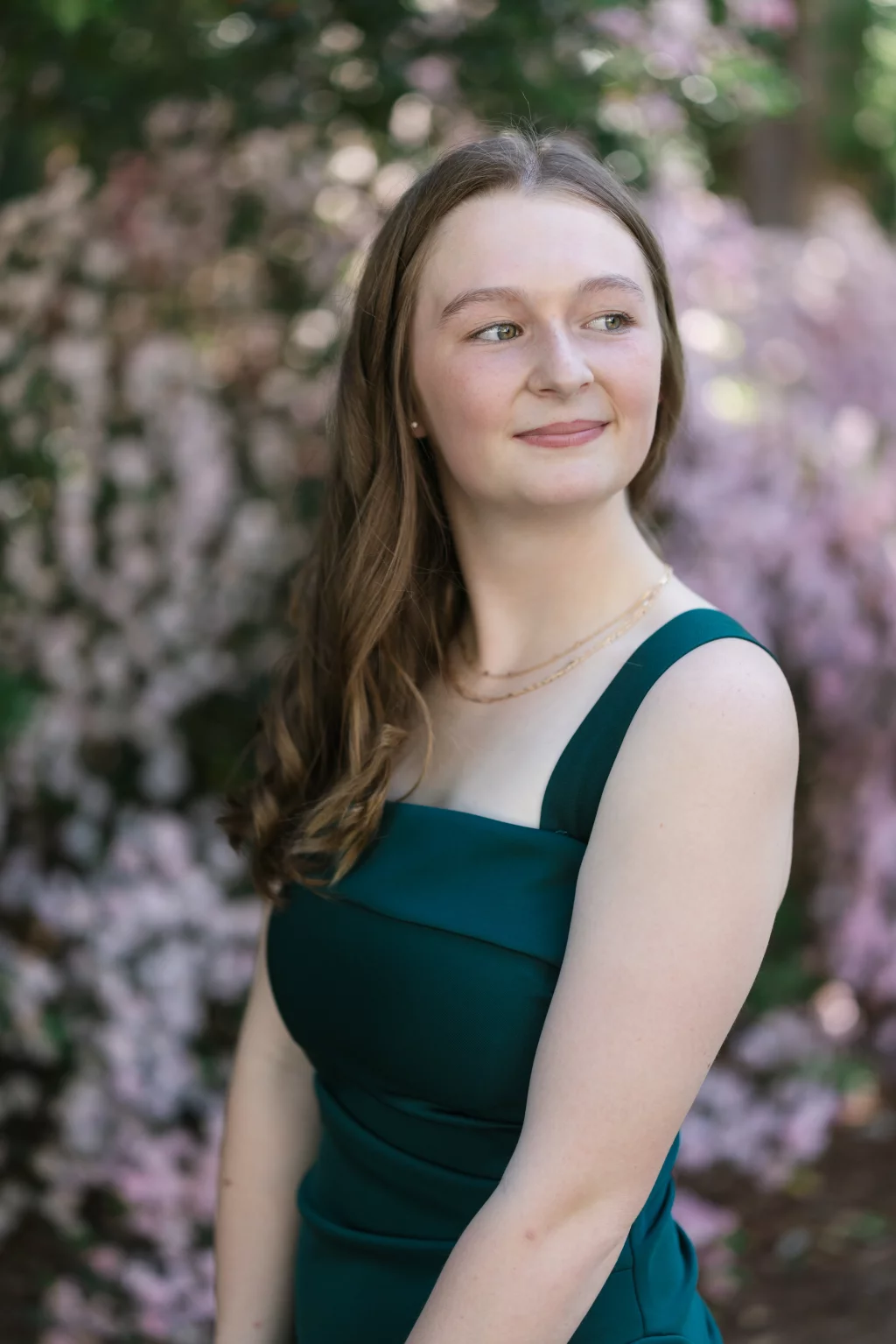 BJA Jr/Sr Banquet Prom at Sue Simpson Park: Young woman with long brown hair in a dark green sleeveless dress, smiling slightly and looking to the side, standing outdoors in front of blurred pink flowers.