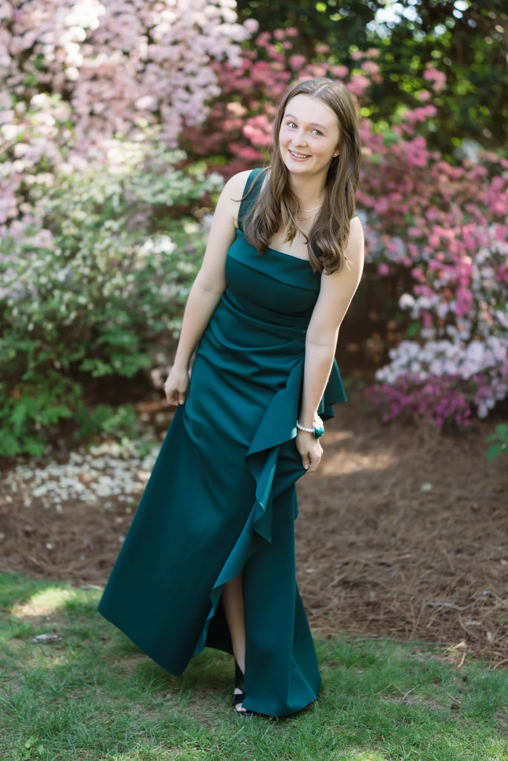 BJA Jr/Sr Banquet Prom at Sue Simpson Park: Young woman in a dark green formal dress standing on grass, smiling, with blooming pink and white flowers in the background.