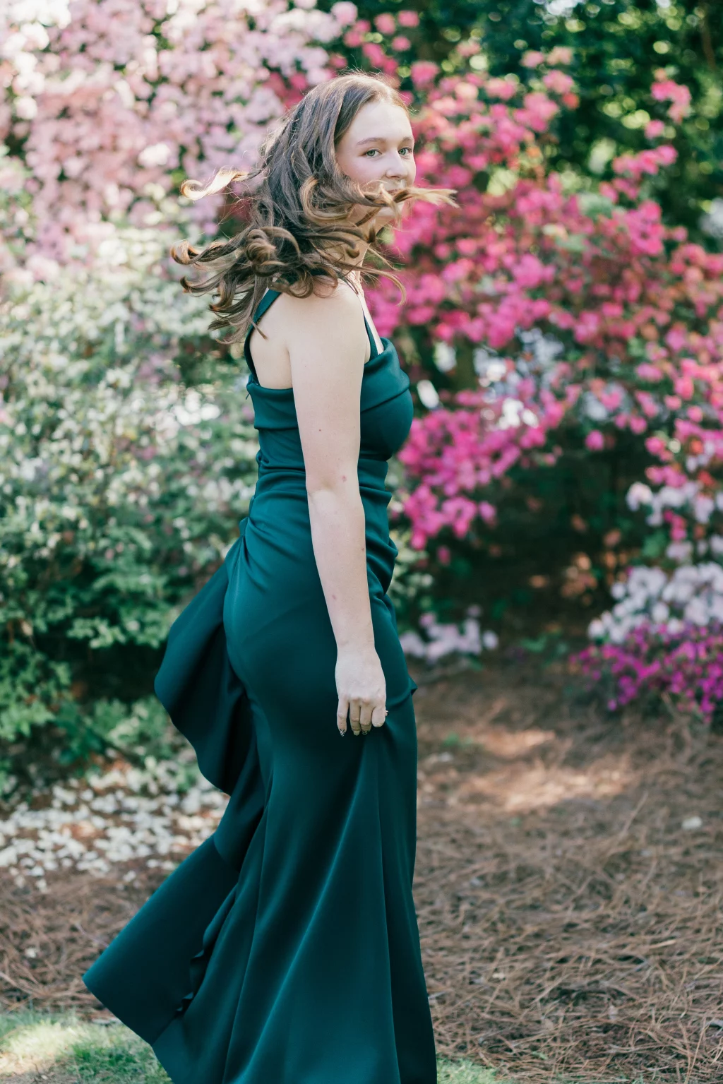 BJA Jr/Sr Banquet Prom at Sue Simpson Park: Young woman in a dark green formal dress turning with flowing hair, standing outdoors in front of blooming pink and white flowers.