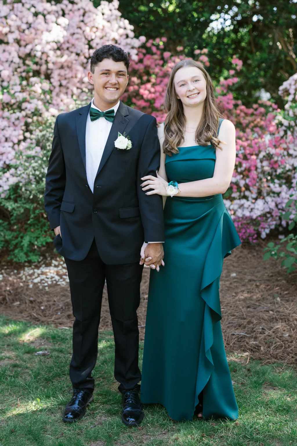 BJA Jr/Sr Banquet Prom at Sue Simpson Park: Two teenagers dressed formally, one in a black suit with a green bow tie and white boutonniere, the other in a sleeveless green gown with a matching corsage, standing arm-in-arm and holding hands in front of blooming pink and white azalea bushes.