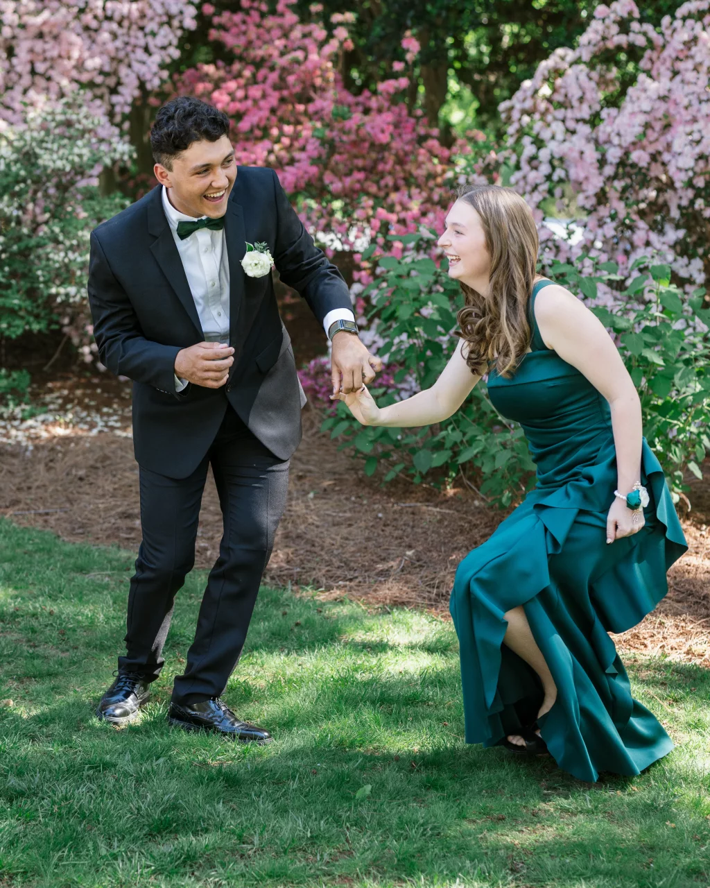 BJA Jr/Sr Banquet Prom at Sue Simpson Park: A young man in a black tuxedo and a young woman in a long teal dress laugh together while holding hands outdoors, surrounded by blooming pink and white flowers.