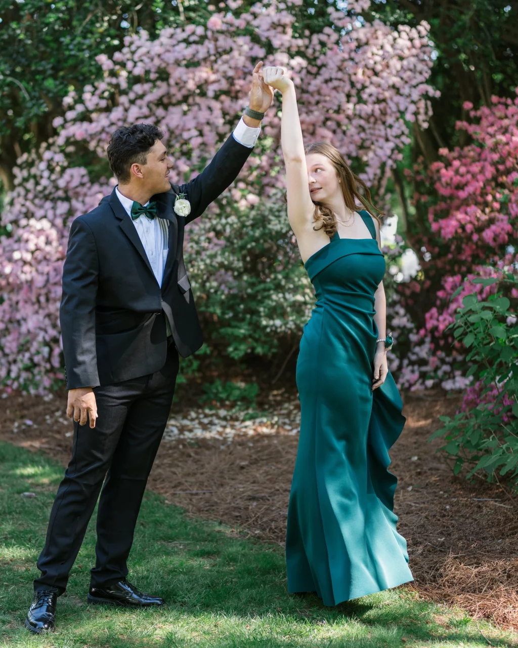 BJA Jr/Sr Banquet Prom at Sue Simpson Park: A young man in a black tuxedo and a young woman in a teal evening gown dance together outdoors, surrounded by blooming pink and purple flowers.