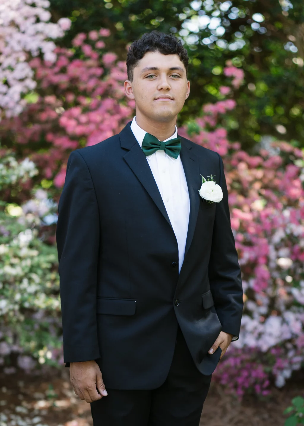 BJA Jr/Sr Banquet Prom at Sue Simpson Park: Young man in a black suit with a green bow tie and white boutonniere, standing in front of blooming pink and white flowers outdoors.