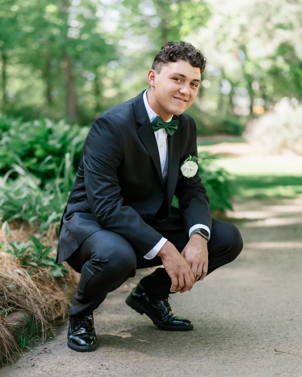 BJA Jr/Sr Banquet Prom at Sue Simpson Park: Young man in a black tuxedo with a green bow tie and white boutonniere squatting on a pathway in a lush, green garden.