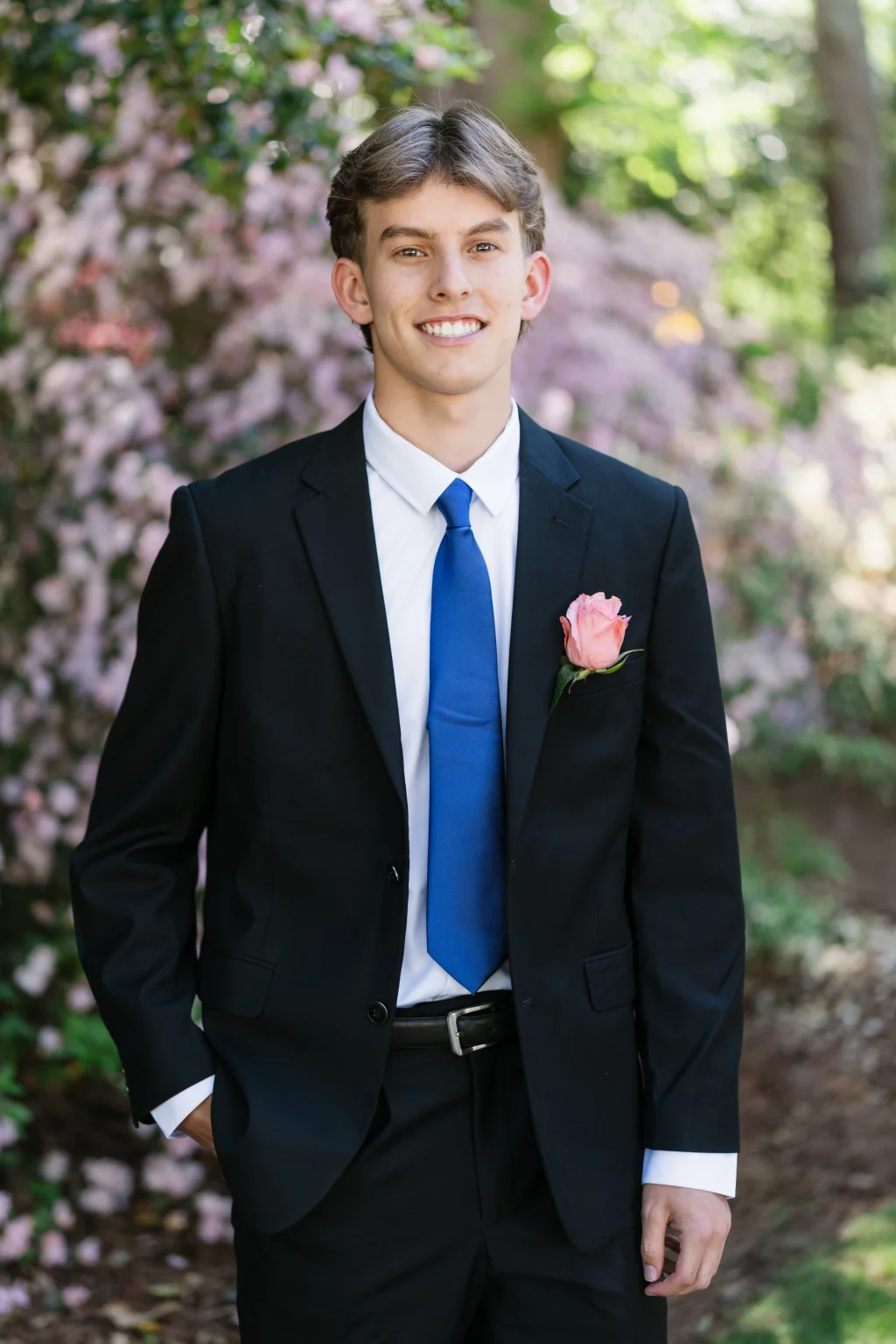 BJA Jr/Sr Banquet Prom at Sue Simpson Park: Young man in a black suit with a blue tie and a pink rose boutonniere, standing outdoors in front of blooming pink flowers.