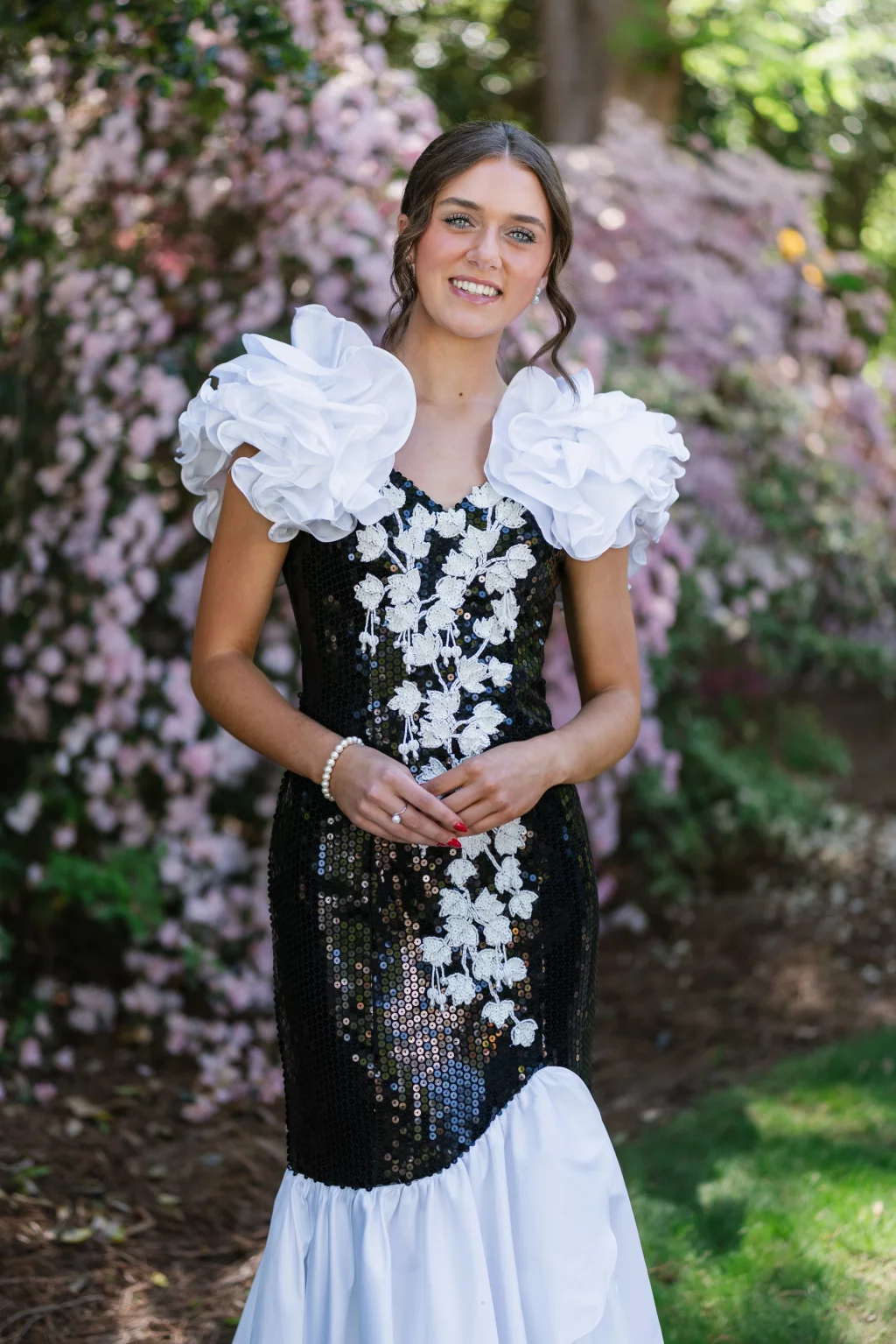 BJA Jr/Sr Banquet Prom at Sue Simpson Park: Young woman standing outdoors in front of pink flowers, wearing a black sequined dress with white floral accents and large ruffled white sleeves, smiling at the camera.