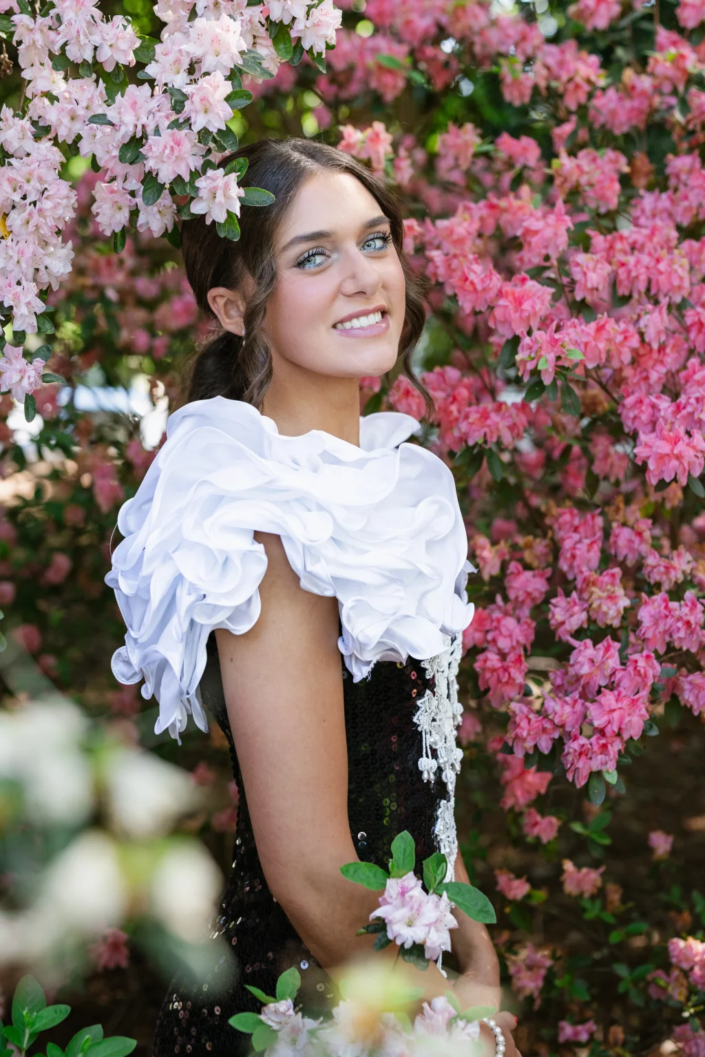 BJA Jr/Sr Banquet Prom at Sue Simpson Park: A young woman with wavy brown hair, wearing a black dress with large white ruffled sleeves, stands smiling among blooming pink and white flowers.