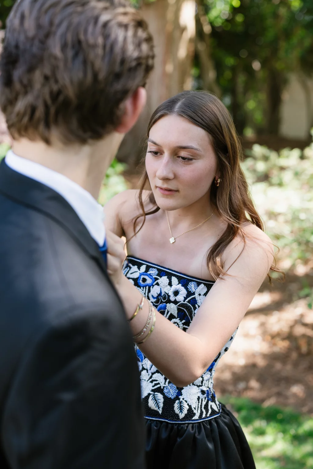 BJA Jr/Sr Banquet Prom at Sue Simpson Park: A young woman in a floral dress adjusts the tie of a young man in a suit outdoors, with greenery in the background.