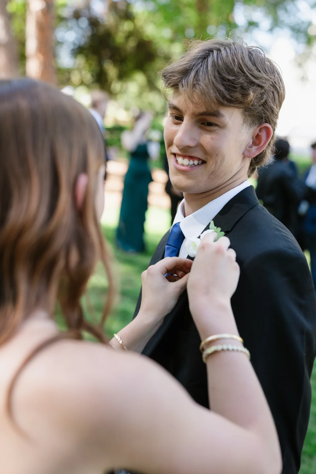 BJA Jr/Sr Banquet Prom at Sue Simpson Park: A young man in a suit smiles as a young woman pins a boutonniere to his lapel outdoors.